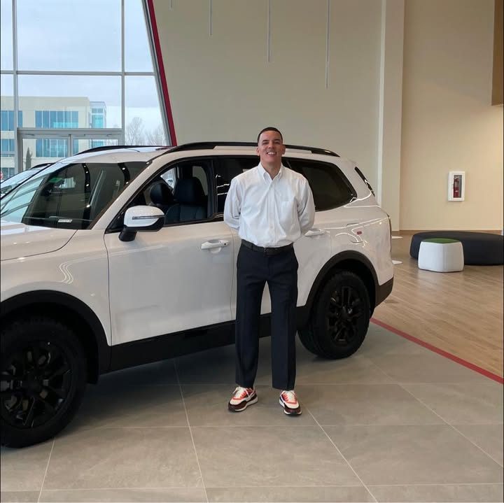 Mario in a white dress shirt, standing beside a white SUV on a dealership showroom floor